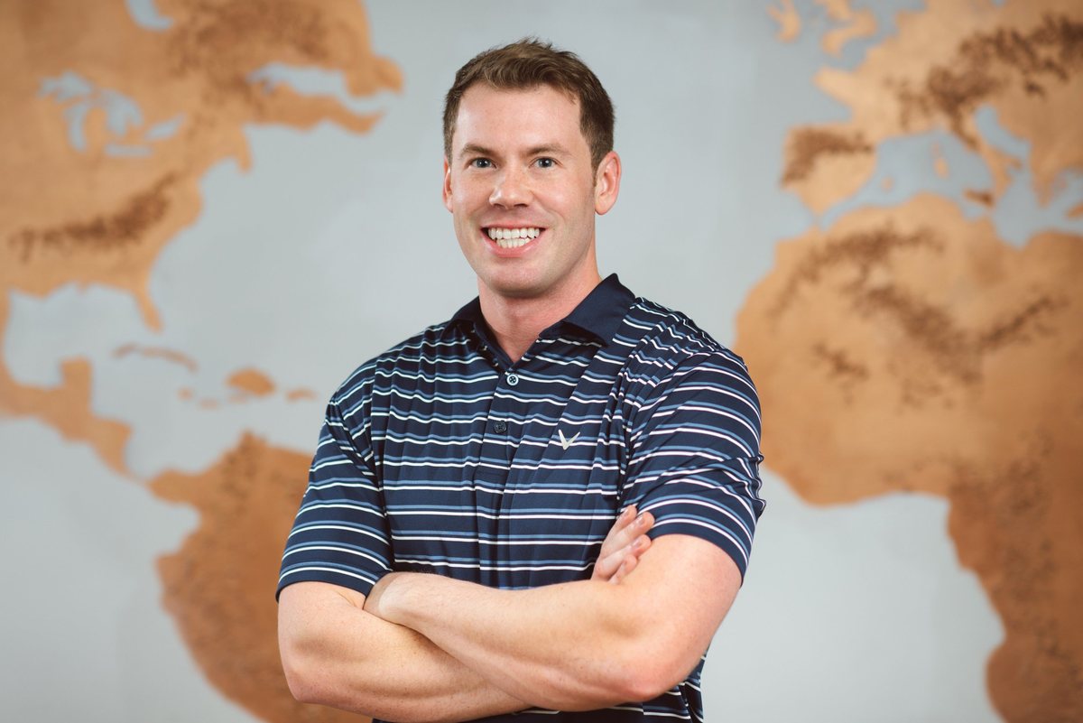 Cameron Clark, arms crossed, smiling in front of a world map